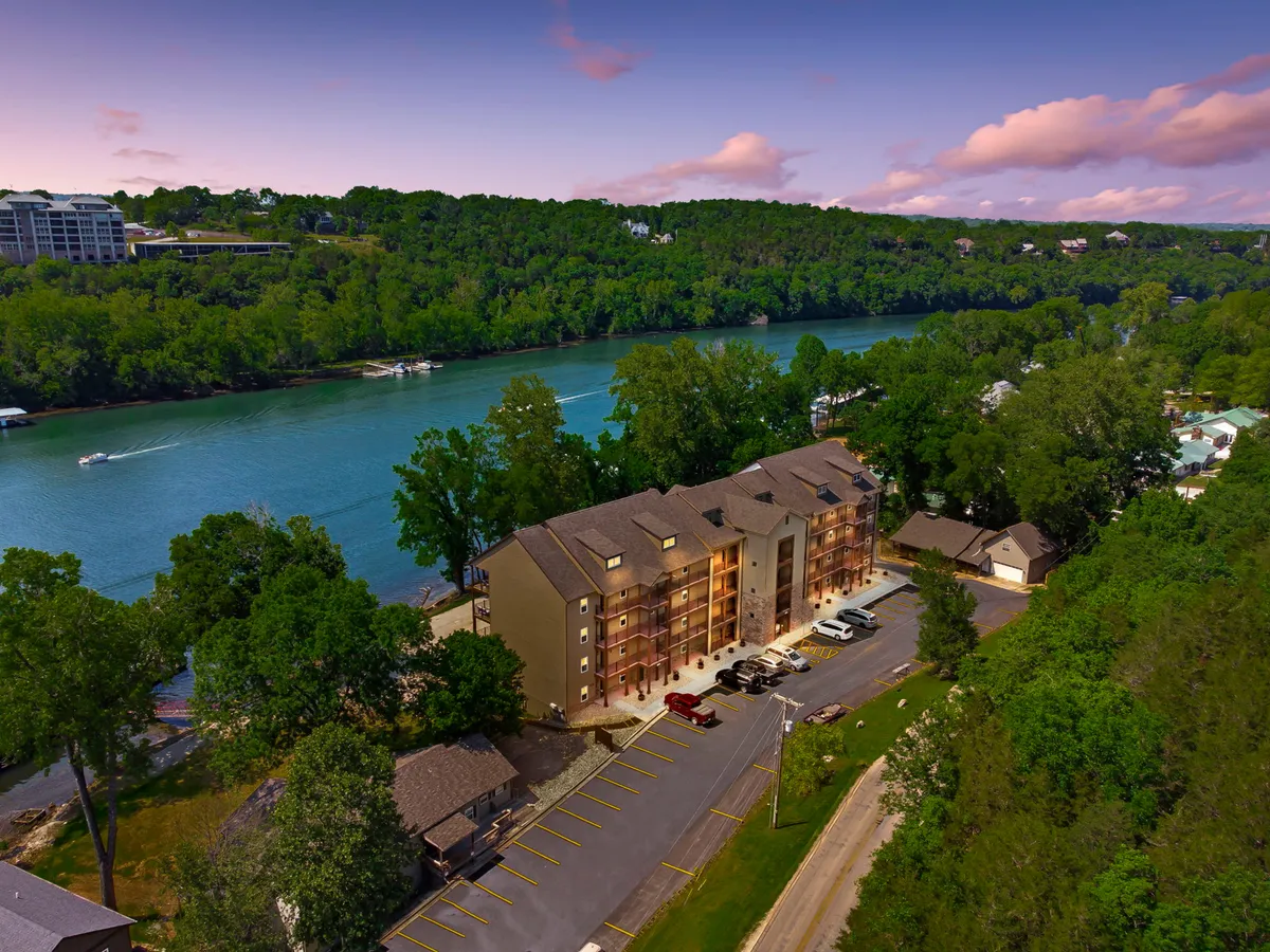 This image shows the exterior of the condo building, beautifully set beside a tranquil river and surrounded by lush trees. The parking area is visible, hinting at easy accessibility, while the inviting river and scenic greenery evoke a sense of relaxation and connection to nature. The scene is set at sunset, adding a warm and welcoming ambiance.