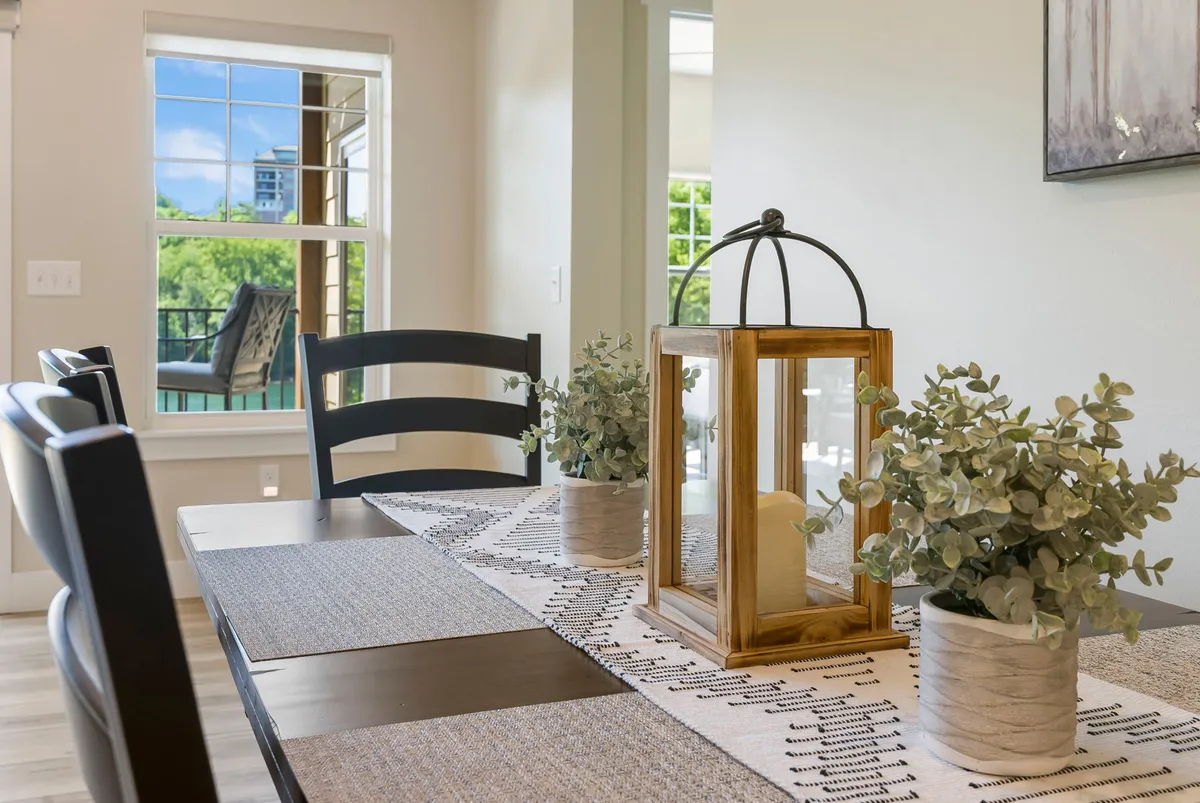 A bright dining area with a large table set for gatherings, adorned with stylish flower arrangements and decorative lanterns, next to expansive windows offering natural light and a calming view.