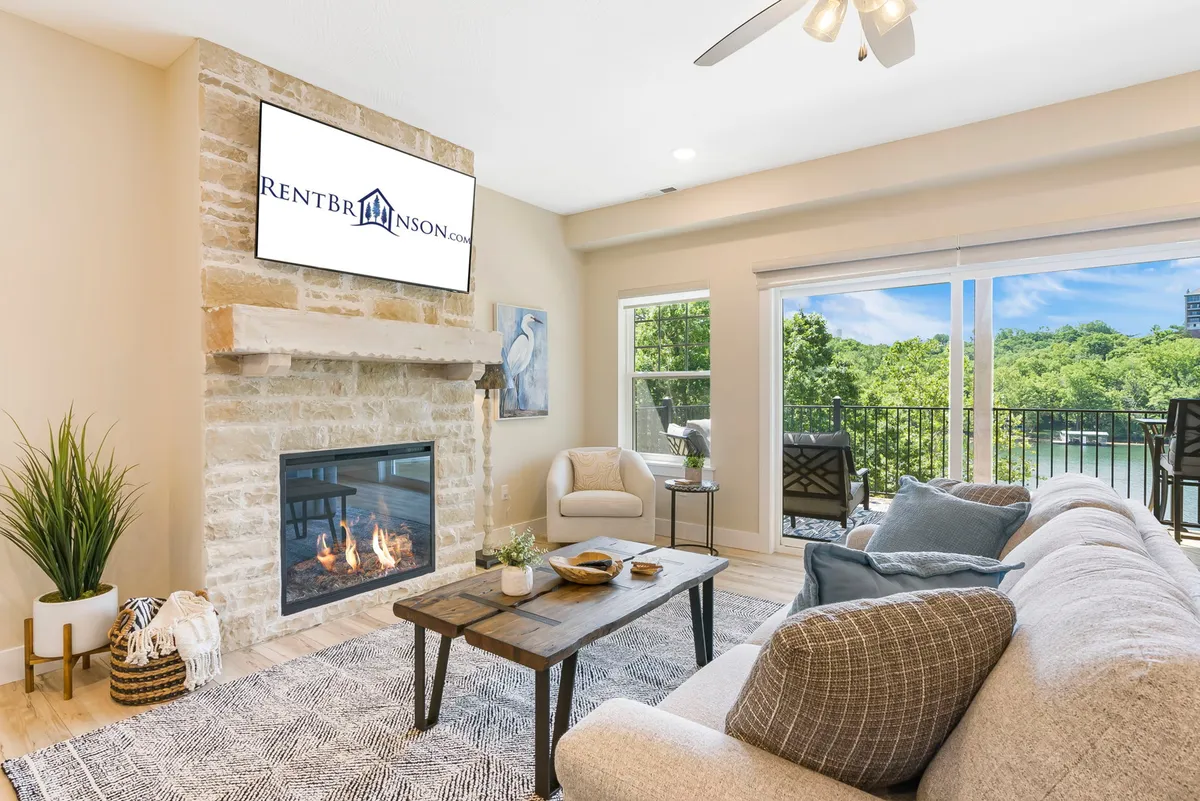 A cozy living room centered around a modern fireplace, with a soft sofa, a coffee table, and a large TV. The room is bathed in natural light from wide windows, with a glimpse of the balcony and outdoor view beyond.