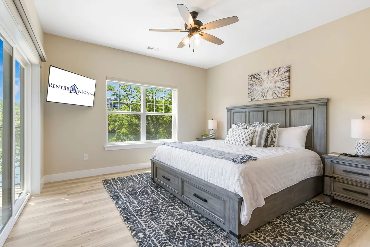 A serene king-sized bedroom featuring a plush bed, large window streaming in natural light, and a television mounted on the wall. The calm, neutral palette and cozy bedding invite relaxation and restful sleep, while the spacious layout underscores a sense of tranquility and privacy.