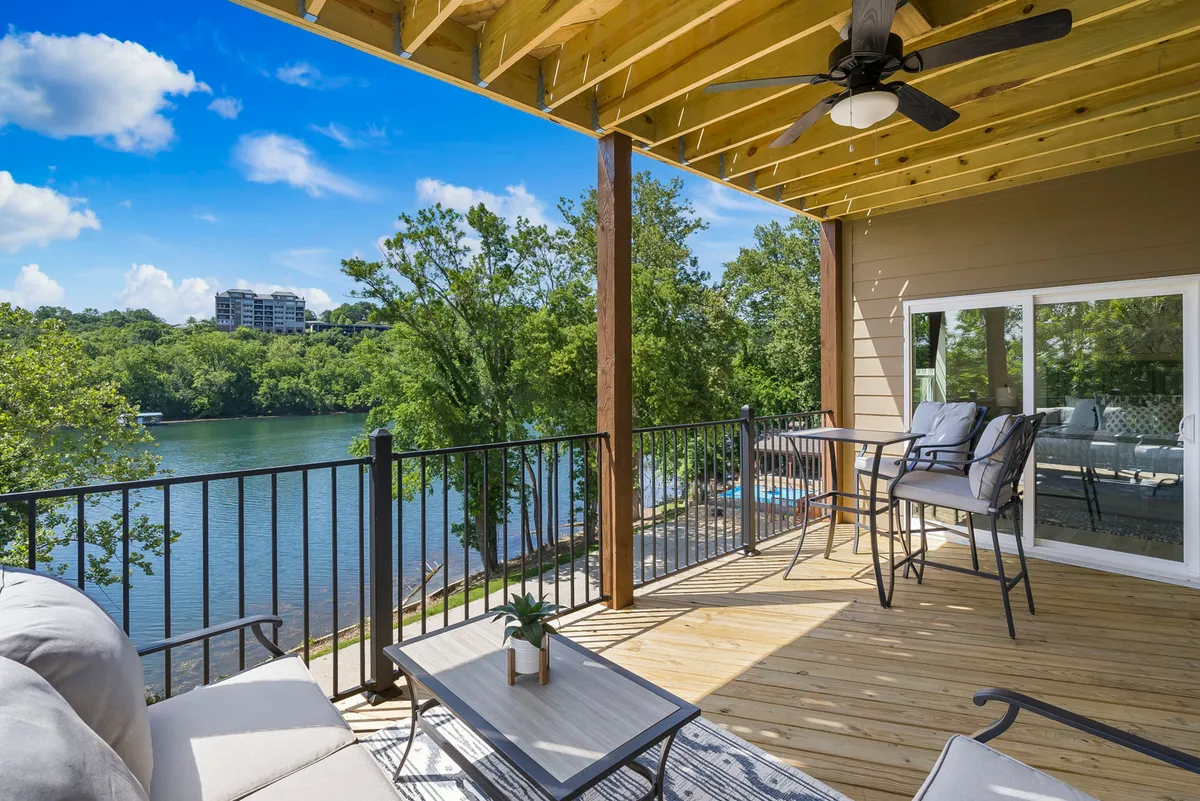 The image features a serene balcony seating area overlooking the shimmering waters of Lake Taneycomo. Comfortable chairs and a decorative plant on the table create an inviting spot to relax, while a breathtaking sunset paints the sky and reflects off the lake, evoking a sense of tranquility and escape.