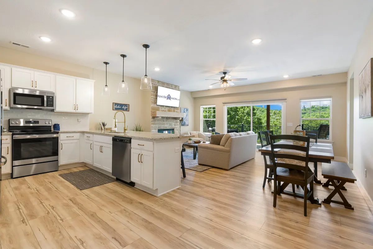 An open-concept space connecting the kitchen, dining, and living areas. The kitchen island and dining table are visible, along with inviting seating and outdoor views through large windows, emphasizing the seamless layout and social possibilities.